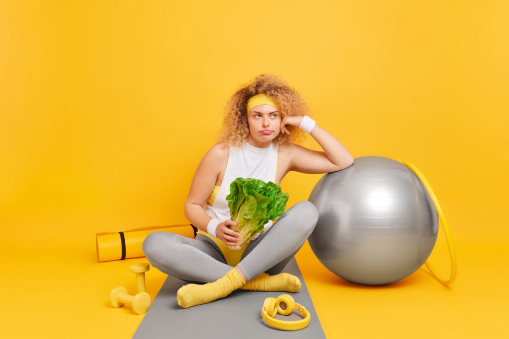 woman with curly hair leans swiss ball holds green vegetable sits crossed legs fitness mat feels tired after training surrounded by fitball hula hoop dumbbells karemat