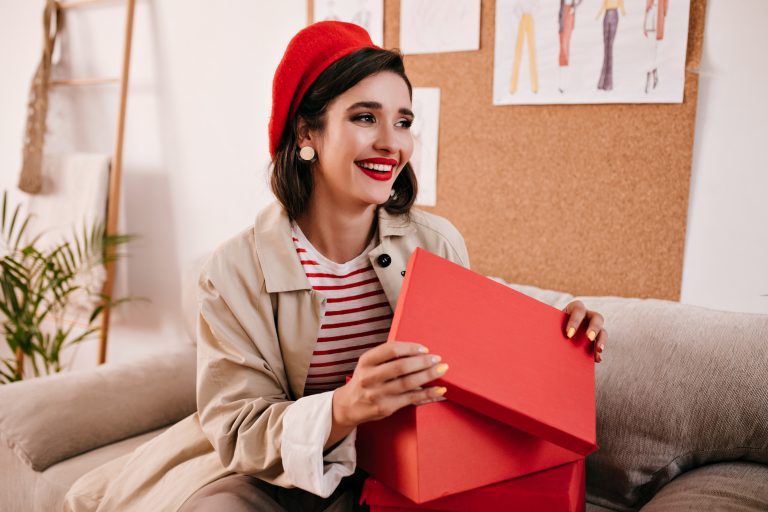 brunette woman in beige trench smiles and opens red box. beautiful lovely girl in striped sweater and bright hat smiles.